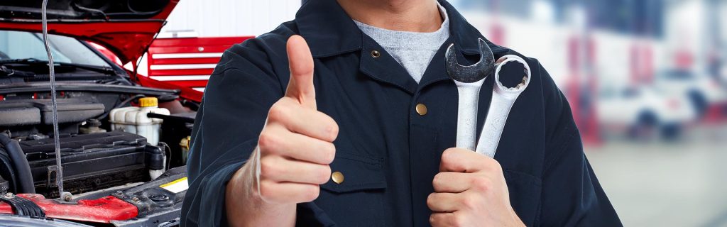 Auto technician holding wrenches and giving a thumbs-up beside an open engine bay.
